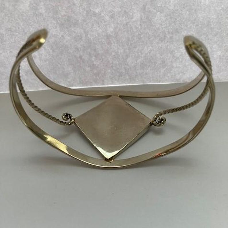 May include: A gold-toned cuff bracelet featuring a central diamond-shaped plaque. The band consists of two parallel wires, with the outer wire having a twisted rope texture. The central plaque is connected to the band by small, coiled metal loops.