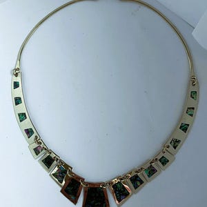 May include: Gold-toned necklace with a curved collar and geometric pendant design. The necklace features rectangular and shield-shaped pendants with iridescent, green, and black inlays. The necklace is displayed on a white background.