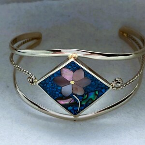 May include: A gold-tone cuff bracelet featuring a central diamond-shaped inlay. The inlay displays a floral motif crafted from iridescent pink shell petals and a blue crushed stone background, accented by a single green leaf-shaped shell piece.