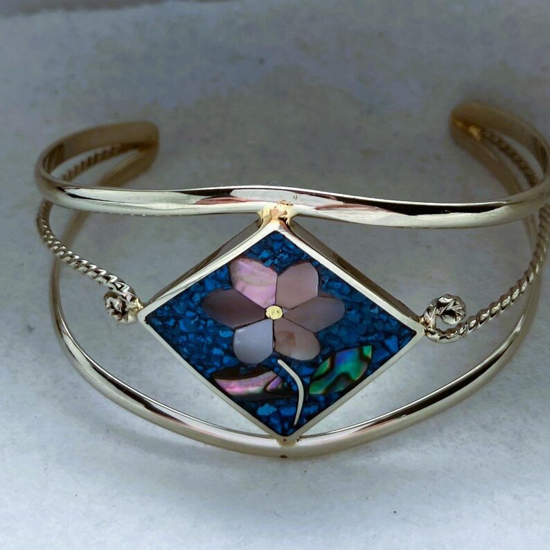 May include: A gold-tone cuff bracelet featuring a central diamond-shaped inlay. The inlay displays a floral motif crafted from iridescent pink shell petals and a blue crushed stone background, accented by a single green leaf-shaped shell piece.
