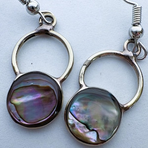 May include: A pair of drop earrings featuring circular silver-toned metal frames. Each earring contains a round, iridescent abalone shell inlay displaying swirling hues of purple, green, and blue. The earrings are attached to silver-toned fishhook wires.