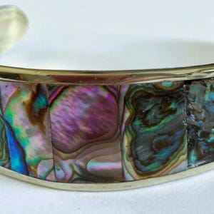 May include: Silver cuff bracelet with iridescent abalone shell inlay. The shell displays a range of colors, including purple, green, and blue. The bracelet has a polished finish and a simple, elegant design.