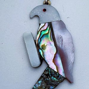 May include: A pendant shaped like a parrot, suspended from a silver-toned chain. The bird features a white head with a small red eye, a body crafted from iridescent abalone shell with green and purple hues, and a dark, textured tail section.
