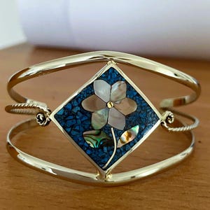 May include: A gold-tone cuff bracelet featuring a central diamond-shaped inlay. The inlay displays a floral motif crafted from iridescent shell pieces set against a background of crushed blue stone, with a small gold-tone dot at the flower center.