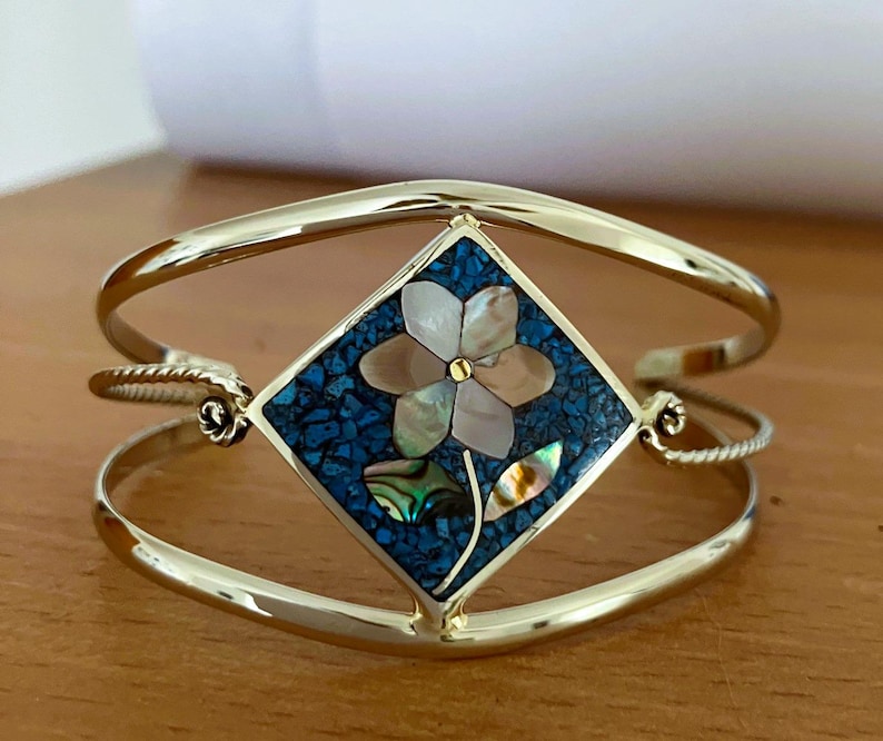 May include: A gold-tone cuff bracelet featuring a central diamond-shaped inlay. The inlay displays a floral motif crafted from iridescent shell pieces set against a background of crushed blue stone, with a small gold-tone dot at the flower center.