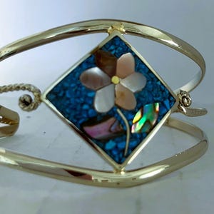 May include: A gold-tone cuff bracelet featuring a central diamond-shaped inlay. The inlay consists of a mosaic of small blue stones surrounding a flower made of iridescent shell petals with a yellow center and a green shell leaf.