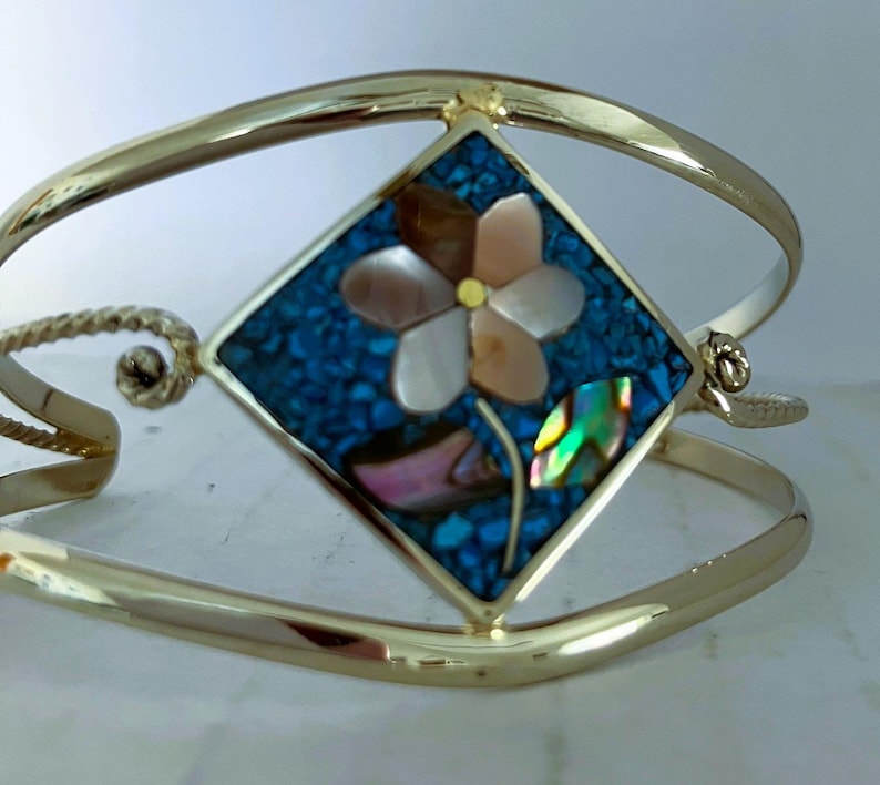 May include: A gold-tone cuff bracelet featuring a central diamond-shaped inlay. The inlay consists of a mosaic of small blue stones surrounding a flower made of iridescent shell petals with a yellow center and a green shell leaf.