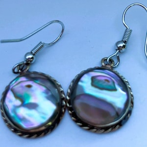 May include: A pair of drop earrings featuring round, iridescent abalone shell cabochons set in twisted metal frames. The earrings have silver-toned metal fishhook wires with a small bead accent, showcasing swirling colors of blue, green, and purple.