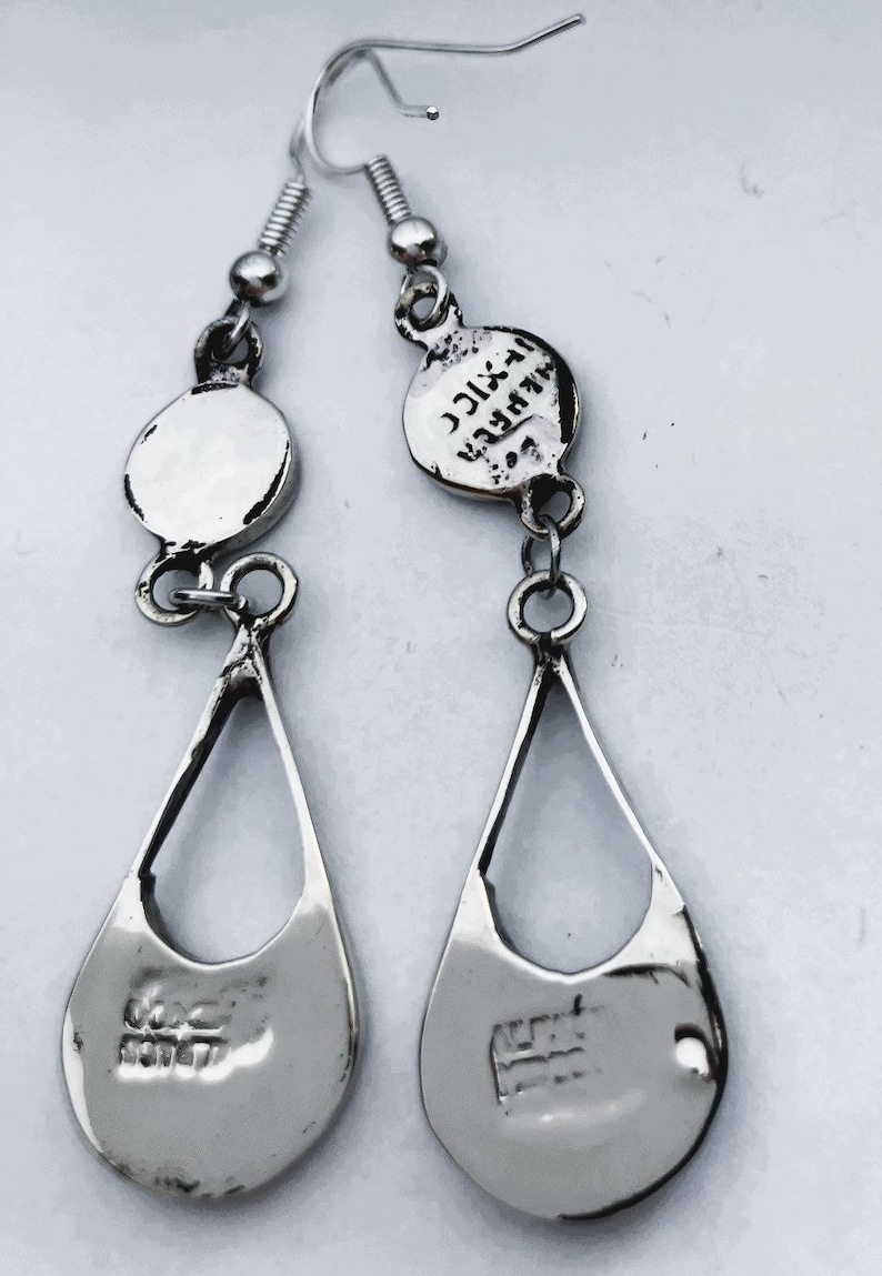 Puede incluir: Un par de pendientes colgantes plateados. Cada pendiente presenta un colgante en forma de l&aacute;grima con un dise&ntilde;o estampado y una pieza superior circular. Los pendientes est&aacute;n conectados por peque&ntilde;os anillos y un gancho.