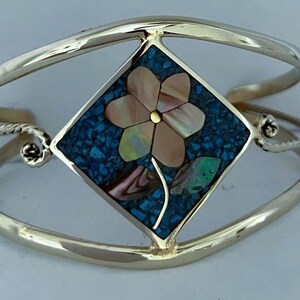 May include: A gold-toned metal cuff bracelet featuring a central diamond-shaped inlay. The inlay displays a flower motif crafted from iridescent shell pieces set against a background of crushed blue stone fragments.