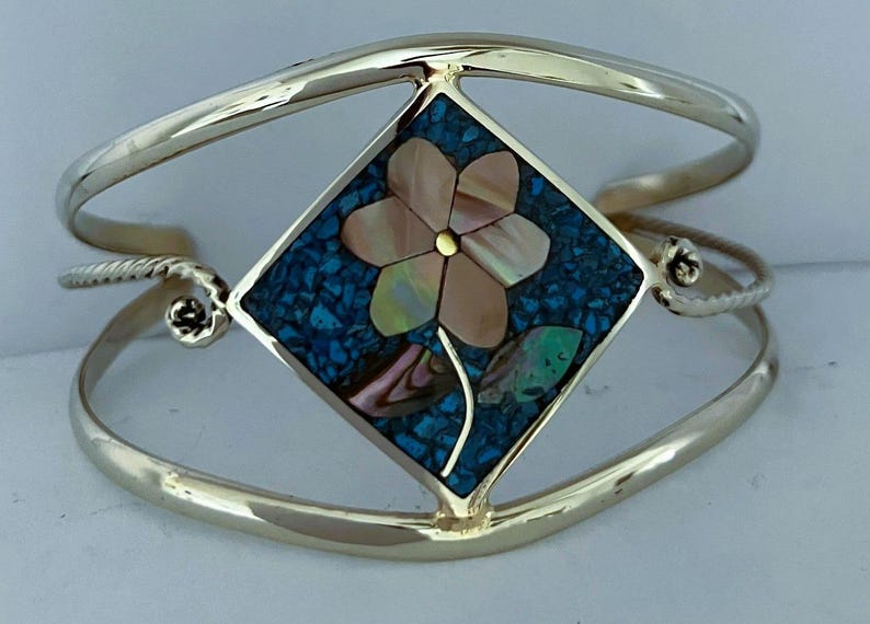 May include: A gold-toned metal cuff bracelet featuring a central diamond-shaped inlay. The inlay displays a flower motif crafted from iridescent shell pieces set against a background of crushed blue stone fragments.