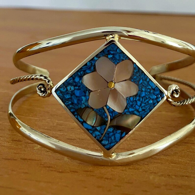May include: A gold-tone cuff bracelet featuring a central diamond-shaped inlay. The inlay displays a flower motif crafted from iridescent shell pieces set against a background of small, crushed blue stone fragments. The metal band has a smooth finish.