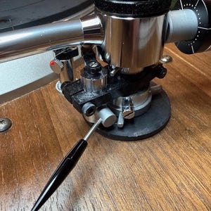 May include: A detailed view of a turntable tonearm. The arm is made of polished chrome, with a black headshell and a black and silver counterweight. The tonearm is set on a wooden surface.