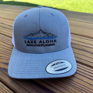 May include: A gray and white snapback hat with the text "LAKE ALOHA DESOLATION WILDERNESS" and a mountain graphic. The hat has a curved brim and a white mesh back. The hat has a "Yupoong Snapback" tag.