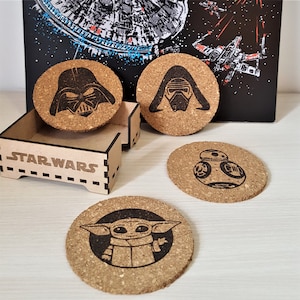 May include: Set of four cork coasters featuring Star Wars characters. The coasters feature Darth Vader, Kylo Ren, BB-8, and Grogu (Baby Yoda). The coasters are in a wooden box with the words "Star Wars" printed on the front.
