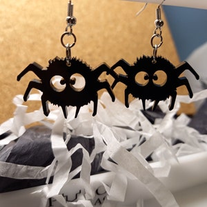 Puede incluir: Un par de pendientes de araña negros con ganchos plateados. Las arañas tienen ojos grandes y redondos y ocho patas.