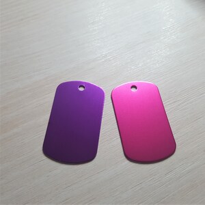 Puede incluir: Dos chapas de perro met&aacute;licas en blanco, una morada y otra rosa. Las chapas tienen una forma rectangular redondeada con un peque&ntilde;o agujero en la parte superior.