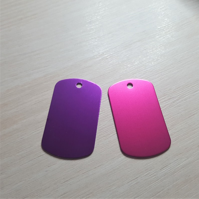 Puede incluir: Dos chapas de perro met&aacute;licas en blanco, una morada y otra rosa. Las chapas tienen una forma rectangular redondeada con un peque&ntilde;o agujero en la parte superior.