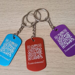 Puede incluir: Tres llaveros con etiquetas met&aacute;licas de diferentes colores. Cada etiqueta tiene un c&oacute;digo QR y el texto "Scansionami!" en blanco. Los llaveros son azul, rojo y morado.