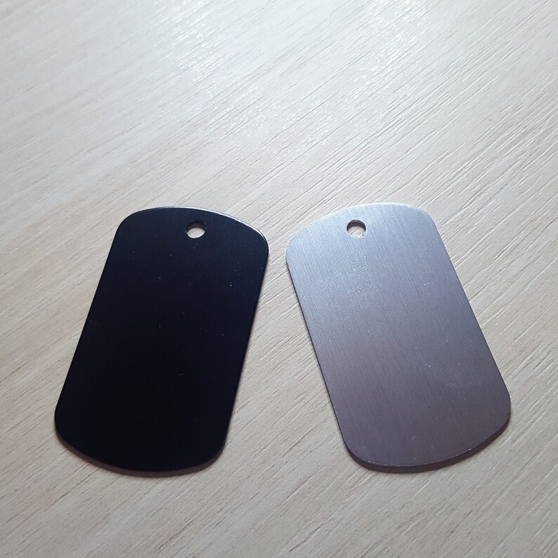 Puede incluir: Dos etiquetas para perros en blanco, una negra y otra plateada, con forma rectangular redondeada y un agujero en la parte superior.