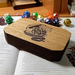 Puede incluir: Una caja de madera con una mancha marrón oscura y un acabado de madera natural. La caja tiene una tapa con bisagras y está grabada con una imagen en blanco y negro de un dragón sosteniendo un dado de 20 caras.