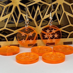 Puede incluir: Pendientes de acrílico naranja con un diseño de estrella de siete puntas. Los pendientes se muestran sobre una superficie de madera con cuatro discos adicionales de acrílico naranja.
