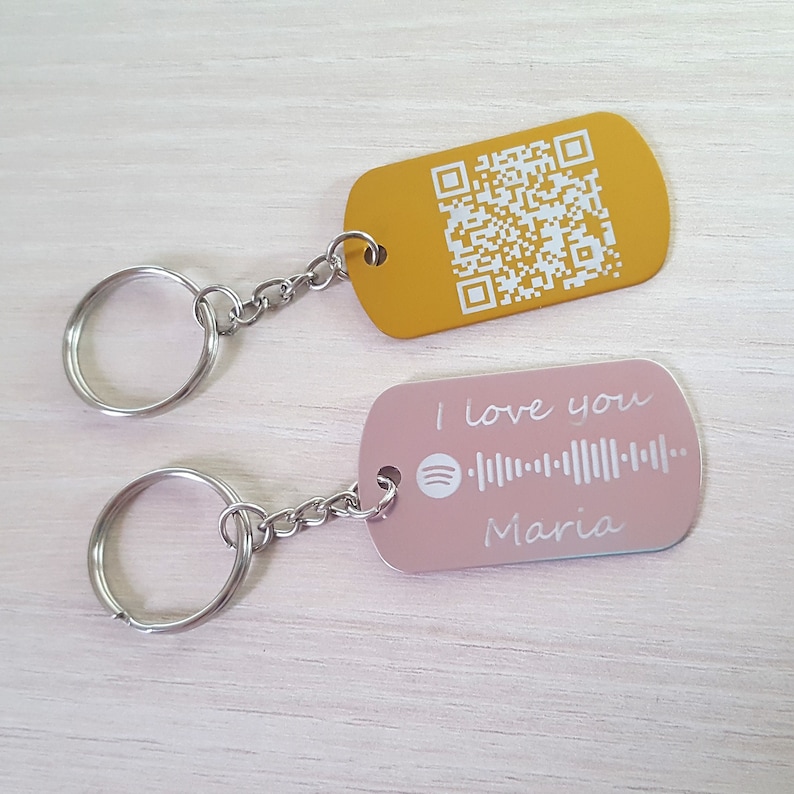 Puede incluir: Dos llaveros con anillos de metal. Un llavero es dorado con un c&oacute;digo QR blanco. El otro llavero es rosa con el texto "Te amo" y un c&oacute;digo de Spotify, seguido del nombre "Maria".