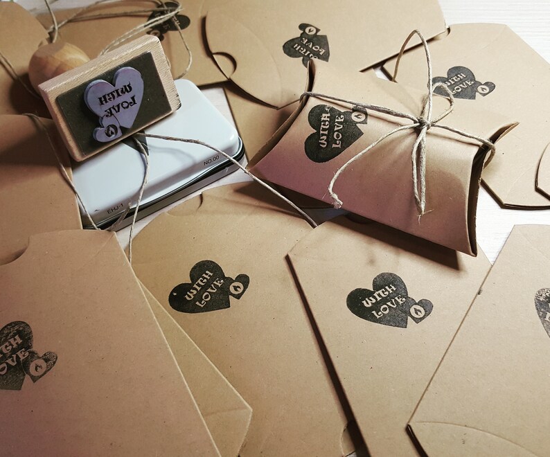 Puede incluir: Una colecci&oacute;n de bolsas de papel marr&oacute;n con un coraz&oacute;n y el texto "Witch Love" estampado en ellas. Las bolsas est&aacute;n atadas con cuerda y algunas est&aacute;n dobladas en forma de almohada. Tambi&eacute;n se ve un sello de madera con el texto "Witch Love".