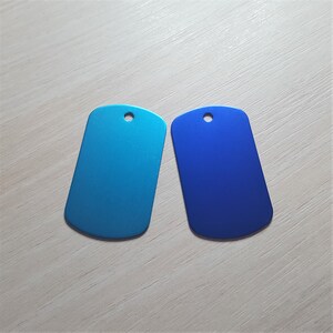 Puede incluir: Dos chapas de perro de metal, una azul y otra turquesa, con forma rectangular redondeada y un peque&ntilde;o agujero en la parte superior.