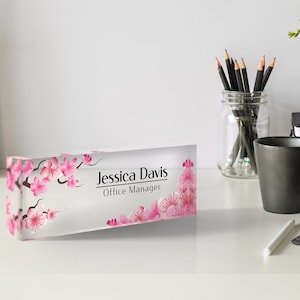 May include: A clear acrylic desk nameplate with a pink cherry blossom design. The nameplate reads "Jessica Davis, Office Manager".
