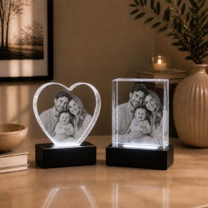 May include: Two clear glass photo blocks with black bases. One block is heart-shaped and the other is rectangular. Both feature a laser-etched black and white photograph of a man, a woman, and a baby. The bases are illuminated from below.