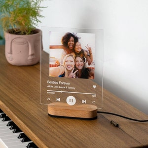 May include: A clear acrylic photo frame with a wooden base. The frame features a photo of four friends and a music player interface with the text "Besties Forever" and the names "Alicia, Jen, Laura & Tammy".