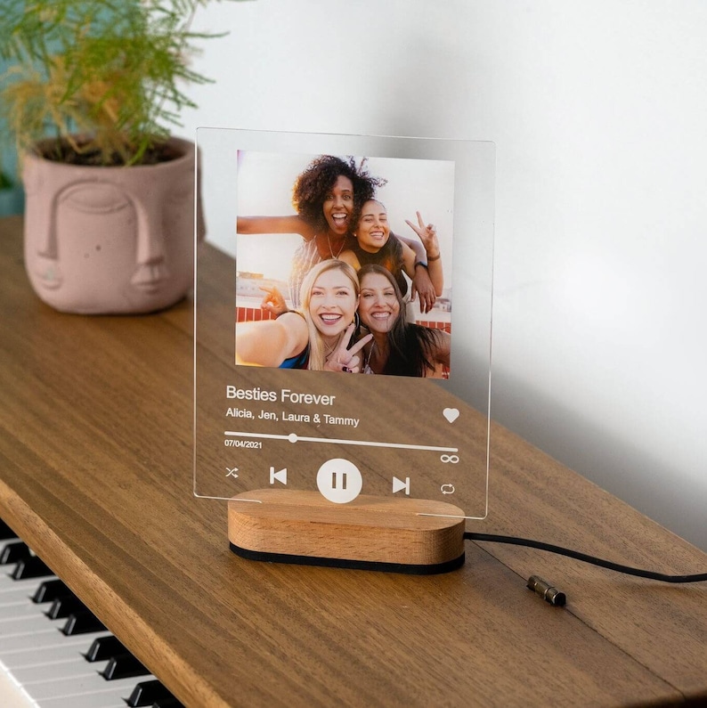 May include: A clear acrylic photo frame with a wooden base. The frame features a photo of four friends and a music player interface with the text "Besties Forever" and the names "Alicia, Jen, Laura & Tammy".