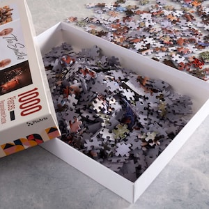 May include: A white box with a 1000 piece personalized photo puzzle inside. The box is open and the puzzle pieces are spilling out. The box has a colorful geometric design and the text "The Smiths" and "1000 Pieces - Personalized Puzzle" on the front. The text "PixArts" is on the bottom right corner of the box.