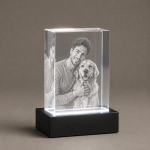 May include: A rectangular clear glass block featuring a laser-etched black and white portrait of a person and a dog. The glass block is mounted on a solid black rectangular base that emits a bright white light from the bottom edge.