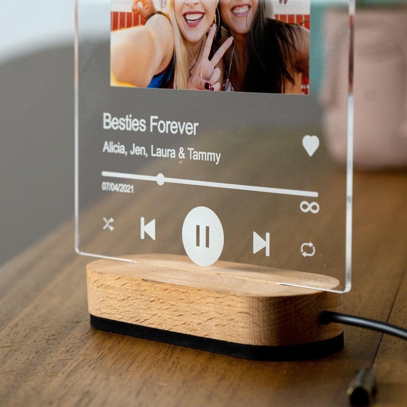 May include: A clear acrylic music player with a photo of two women smiling and making peace signs. The text "Besties Forever" is printed on the player, along with the names "Alicia, Jen, Laura & Tammy" and the date "07/04/2021". The player has a wooden base and a black cord.