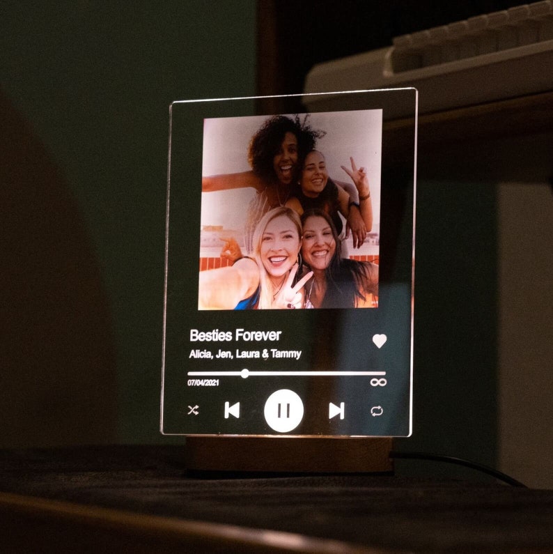 May include: A clear acrylic photo frame with a lit-up music player design. The frame displays a photo of four friends smiling and making peace signs. The music player features a heart icon, a play button, and a date: 07/04/2021. The text "Besties Forever" and "Alicia, Jen, Laura & Tammy" are displayed below the photo.