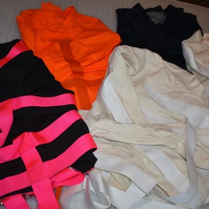 Puede incluir: Una colección de prendas de vestir en varios colores. Hay una parte superior negra con rayas rosas brillantes, una parte superior naranja, una parte superior azul marino y una prenda de color crema con rayas blancas. Los artículos están dispuestos sobre una superficie de color claro.