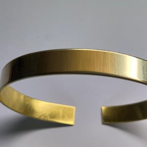 Puede incluir: Un brazalete simple de metal dorado. El brazalete es liso y tiene un acabado pulido.