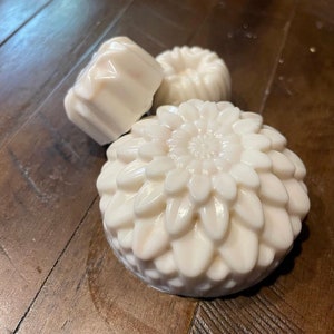 May include: Three white soaps of varying shapes rest on a wooden surface. One soap is a large, round, flower-shaped design with detailed petals. Another is a small, round, ridged shape, and the third is a rectangular shape with a ridged design.