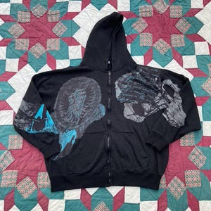 May include: Black zip-up hoodie with a hood and front pockets. The hoodie features large, abstract graphic designs in shades of gray and blue on the front and sleeves. The hoodie is displayed on a patterned quilt.