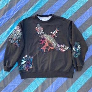May include: Black sweatshirt with a graphic print of a winged eagle and the word "Eternal". The eagle has a red, white, and blue design. The sweatshirt has a crew neck and long sleeves.