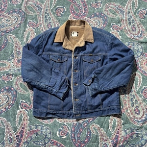 May include: A blue denim jacket featuring a tan corduroy collar and a tan faux-fur lining. The jacket includes two chest pockets with button-down flaps, a front button closure, and long sleeves with buttoned cuffs. The denim has a classic wash.