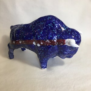 May include: A blue ceramic sculpture of a bison with white and brown accents. The bison is standing on all four legs and has a speckled blue glaze.