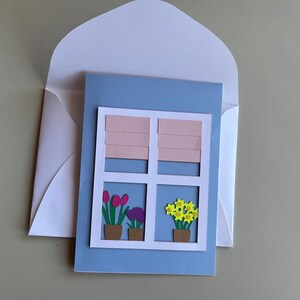 May include: A handmade greeting card featuring a window design with pink blinds and potted flowers. The card is light blue with a white frame, and the flowers include tulips and yellow blooms. The card is inside a white envelope.
