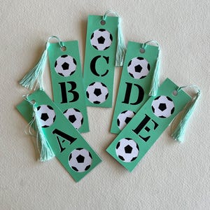 May include: Five rectangular mint green bookmarks, each featuring a black and white soccer ball graphic and a single black letter. The letters A, B, C, D, and E are displayed. Each bookmark includes a hole at the top with a mint green tassel.