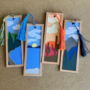 May include: Four landscape-themed bookmarks with colourful layered paper designs. Each bookmark features a different scene: a mountain range, a sunset with cacti, a lake with a sun, and a forest. Each has a tassel.