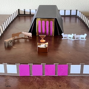 Puede incluir: Maqueta de una estructura religiosa, con techo negro, detalles dorados y paneles verticales rosas. El modelo incluye pequeñas figuras de animales, una copa dorada y una estructura rectangular. El perímetro está rodeado por una valla blanca y negra.