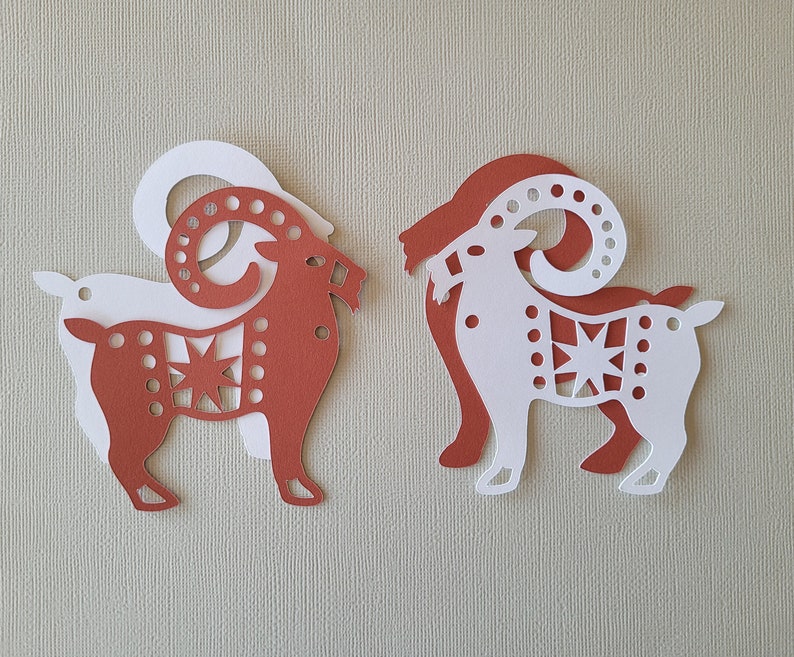 May include: Two paper cutouts of goats, one red and one white, with intricate designs. The goats are facing each other and have a star design on their bodies.