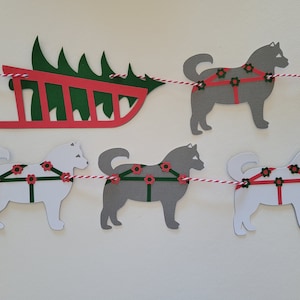 May include: A red sleigh with a green Christmas tree on top, pulled by four white and grey dogs. The dogs are wearing harnesses with red flowers and green straps. The sleigh and dogs are part of a paper garland.